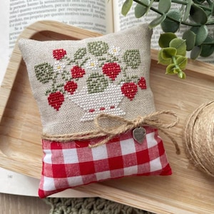 May include: A small, rectangular pillow with a cross-stitched strawberry design. The top section is beige, and the bottom is red and white checkered fabric. A jute bow and a heart-shaped charm that says "Made with Love" are attached.