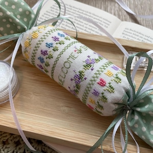 May include: A handcrafted fabric cylinder with floral embroidery and the word "WELCOME" is presented on a wooden tray. It's adorned with ribbons and polka-dot fabric details. A small jar containing a white substance is also on the tray.