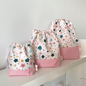 Pode incluir: Três sacos com cordão com um padrão floral em rosa, verde, amarelo e branco. Os sacos têm uma base rosa e um cordão branco. Os sacos são de tamanhos diferentes e são exibidos numa prateleira branca.