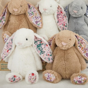 May include: Five plush bunny rabbits in shades of tan, white, grey, and brown. The bunnies have long ears with floral patterned fabric on the inside. One bunny has the name "Clara" embroidered on its ear. The bunnies are sitting on a surface.