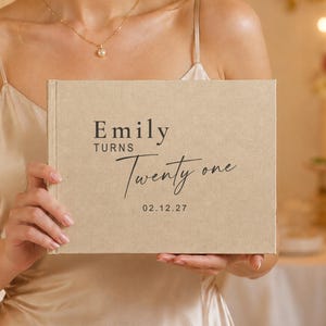 May include: A rectangular beige fabric-covered photo album with black printed text that reads Emily TURNS Twenty one and the date 02.12.27. The cover has a textured linen-like appearance and a clean, centered layout with a mix of serif and script fonts.