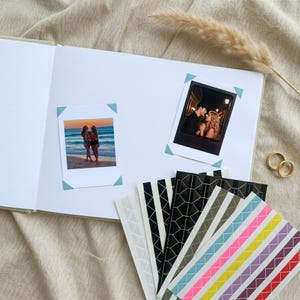 以下が含まれることがあります： 白いスクラップブックに2枚の正方形の写真が貼られており、1枚は夕暮れのビーチにいる2人、もう1枚は夜の2人を写しています。様々な色や模様の粘着式フォトコーナーシールが扇状に並べられています。