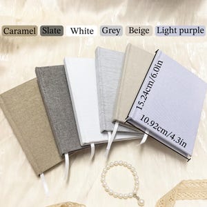 Op de afbeelding: Zes met stof beklede notitieboeken in karamel, leisteen, wit, grijs, beige en lichtpaars. Elk notitieboek heeft een wit lint als bladwijzer. Het lichtpaarse notitieboek is voorzien van de afmetingen 15,24 cm bij 10,92 cm.
