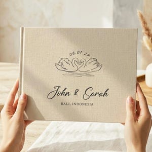 May include: A beige linen-covered photo album featuring a black line art illustration of two swans facing each other. The text reads 06.01.27, John & Sarah, and BALI, INDONESIA, printed in a dark font centred on the front cover of the book.