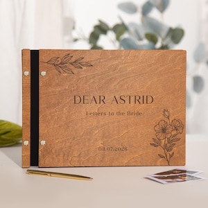 May include: A wooden guest book with the text "DEAR ASTRID Letters to the Bride" and the date 03.07.2026. The cover features engraved floral and leaf designs. A black spine with silver rivets holds the pages together. A gold pen and photos are nearby.