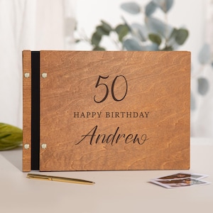 Puede incluir: Un álbum de fotos rectangular de madera con un acabado de grano de madera marrón natural. La cubierta presenta el texto grabado 50 HAPPY BIRTHDAY Andrew en una mezcla de fuentes serif y script. El álbum está encuadernado con tela negra y remaches.