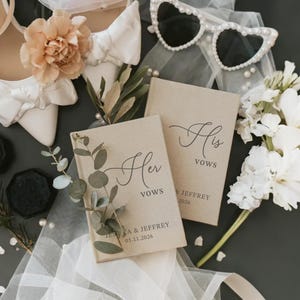 Op de afbeelding: Twee beige gelofteboeken met zwarte tekst Her Vows en His Vows, gepersonaliseerd met Jessica & Jeffrey 05.11.2026. Omringd door witte hakken met strikken, parel-omrande hartvormige zonnebrillen, eucalyptus takjes en witte bloemstukken.