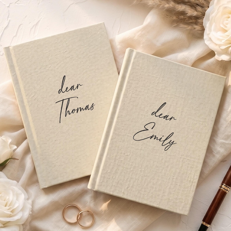 Op de afbeelding: Twee cr&egrave;mekleurige hardcover notitieboeken met een textuur van stof. Het linker notitieboek heeft de zwarte opdruk "dear Thomas" en het rechter notitieboek heeft de zwarte opdruk "dear Emily" in een cursief lettertype.