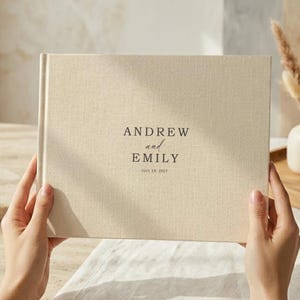May include: A landscape-oriented photo album with a textured beige fabric cover. The front features dark brown printed text that reads ANDREW and EMILY with the date JULY, 19, 2027 centered below the names in a clean, minimalist serif font.