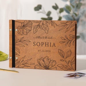 May include: A rectangular wooden photo album or guestbook with a dark brown finish. The cover features laser-engraved floral illustrations, the text "letters to the bride", the name "SOPHIA", and the date "07.12.2026" in a dark brown font.