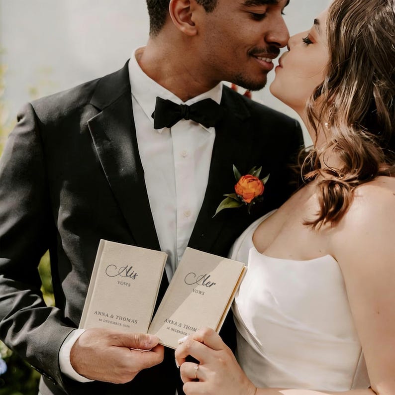 Op de afbeelding: Twee beige gelofteboekjes met zwarte tekst die His Vows en Her Vows leest, gepersonaliseerd met de namen Anna & Thomas en de datum 10 december 2024. De boekjes worden vastgehouden door twee personen in formele trouwkleding, een pak en een jurk.
