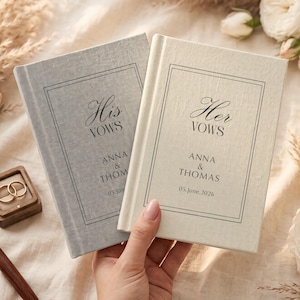 Op de afbeelding: Twee hardcover gelofteboeken met stoffen kaft, &eacute;&eacute;n grijs en &eacute;&eacute;n cr&egrave;me. Het grijze boek is getiteld His Vows en het cr&egrave;mekleurige boek Her Vows. Beide bevatten de namen Anna & Thomas en de datum 05.June.2026 gedrukt in zwarte inkt.