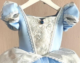 May include: A light blue princess-style dress with a tulle skirt. The bodice features a white lace panel and a jewel accent. The sleeves and skirt are trimmed with silver lace. The dress is hanging on a white hanger.