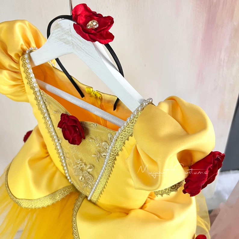 May include: Yellow satin dress with a tulle skirt and red rose accents. The dress has a lace trim and a zipper closure in the back. The dress is on a white hanger.