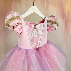 May include: A pink princess dress with a square neckline and short, puffed sleeves. The bodice features white lace detailing and a jewel accent. The skirt is layered with pink and lavender tulle, creating a shimmering effect. Gold trim accents the sleeves.