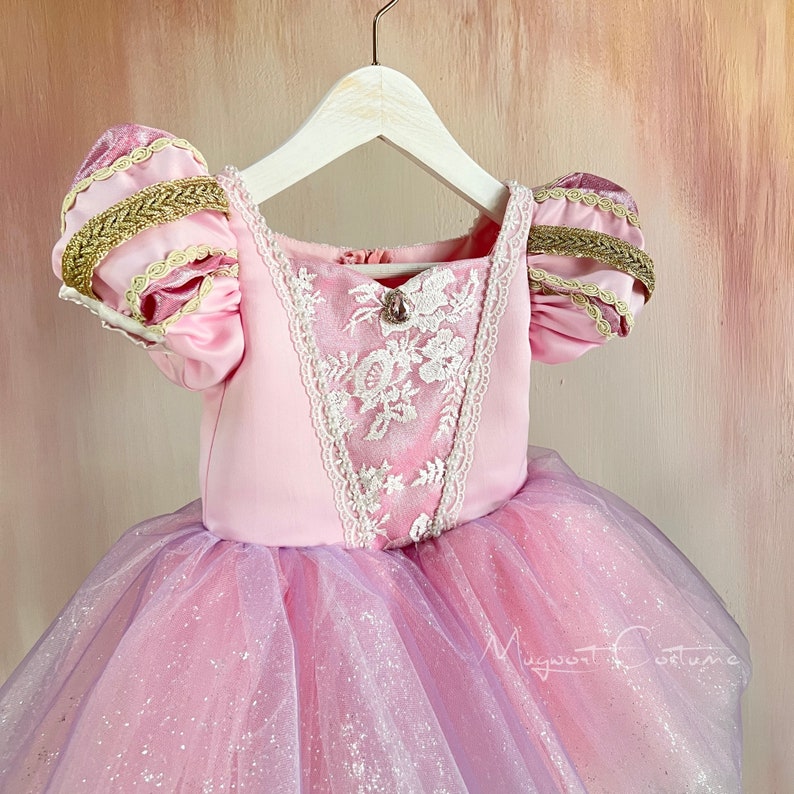 May include: A pink princess dress with a square neckline and short, puffed sleeves. The bodice features white lace detailing and a jewel accent. The skirt is layered with pink and lavender tulle, creating a shimmering effect. Gold trim accents the sleeves.