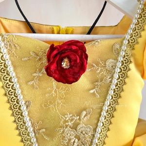 May include: A yellow dress with gold embroidery and a red rose embellishment. The dress has a white pearl trim and a black ribbon.