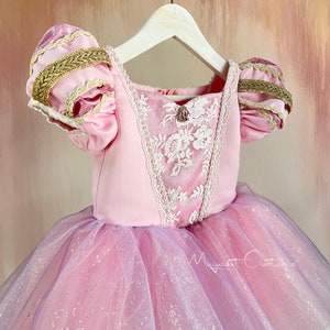 May include: A pink princess dress with a bodice featuring white lace detailing and a jewel accent. The sleeves have gold trim and the skirt is layered with pink and lavender tulle. The dress is hanging on a white hanger.