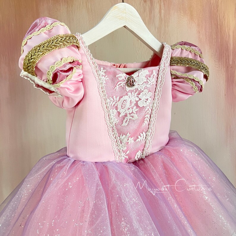 May include: A pink princess dress with a bodice featuring white lace detailing and a jewel accent. The sleeves have gold trim and the skirt is layered with pink and lavender tulle. The dress is hanging on a white hanger.
