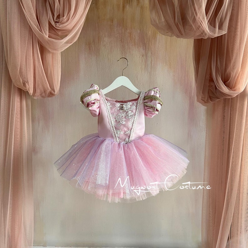 May include: A pink tulle tutu with a pink satin bodice and white lace trim. The bodice has short sleeves with gold trim. The tutu is hanging on a white hanger against a pink background.