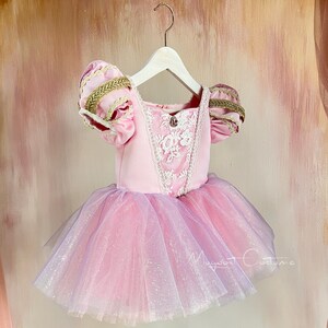 May include: A pink and lavender princess costume dress with a tulle skirt. The bodice has lace detailing and gold trim on the sleeves. The dress is hanging on a white hanger.