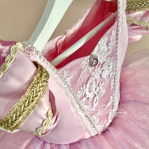 May include: Pink princess costume with a white lace bodice, gold trim, and a pearl-like trim. The bodice has a pink button with a rhinestone in the center.