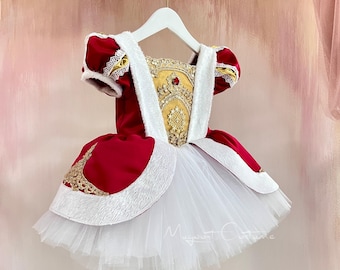 May include: A red and white dress with gold accents, hanging on a white hanger. The dress features a white tulle skirt, red bodice with gold embroidery, and white faux fur trim. The dress has short sleeves and a decorative bodice.