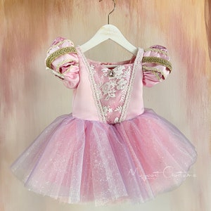 May include: A pink and lavender princess-style dress with a tulle skirt and puff sleeves. The bodice features white lace detailing and a small jewel. The sleeves are accented with gold trim. The dress is hanging on a white hanger.