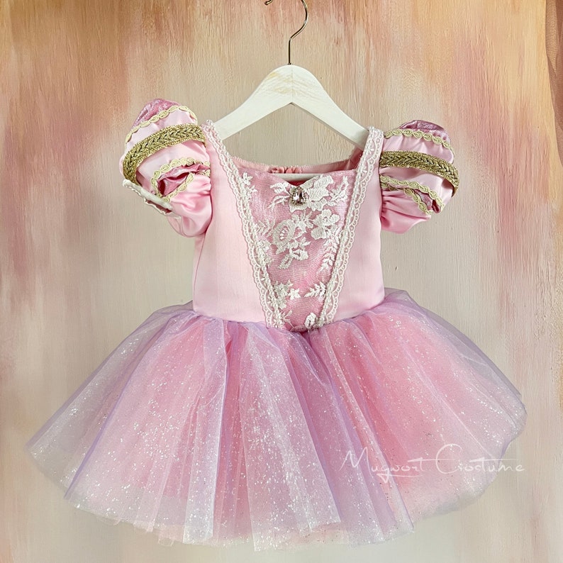 May include: A pink and lavender princess-style dress with a tulle skirt and puff sleeves. The bodice features white lace detailing and a small jewel. The sleeves are accented with gold trim. The dress is hanging on a white hanger.