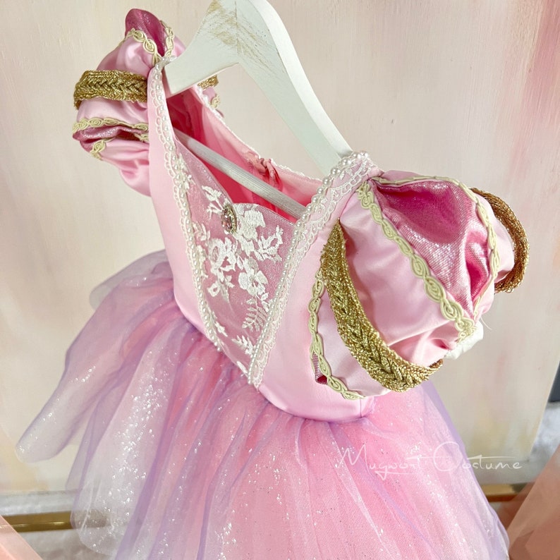 May include: A pink and purple princess dress with a tulle skirt, gold trim, and white floral embroidery. The dress is hanging on a white hanger. The bodice has a jewel accent.
