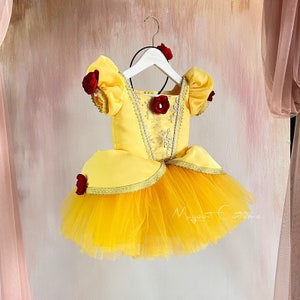 May include: Yellow tulle and satin dress with red rose accents. The dress has a fitted bodice with a sweetheart neckline and a full skirt. The dress is on a white hanger.