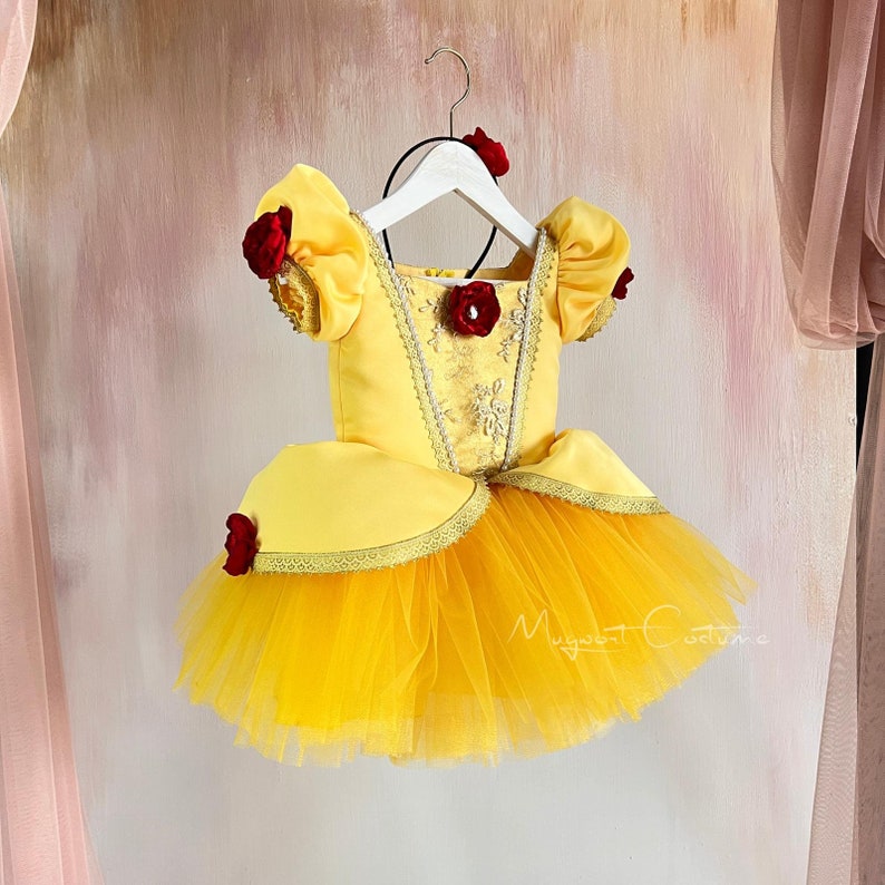 May include: Yellow tulle and satin dress with red rose accents. The dress has a fitted bodice with a sweetheart neckline and a full skirt. The dress is on a white hanger.