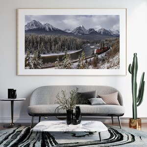 May include: A framed photograph of a red train traveling through a snowy mountain landscape. The train is traveling along a river and through a forest. The mountains are covered in snow and the sky is cloudy.