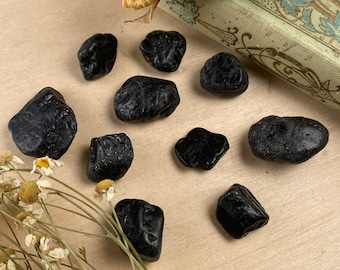 May include: A collection of ten black obsidian tumbled stones on a light brown surface. The stones are various sizes and shapes.