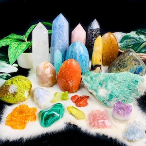 May include: A collection of colorful and polished crystals and stones, including green, blue, red, orange, and white. The stones are arranged on a white, fluffy surface.