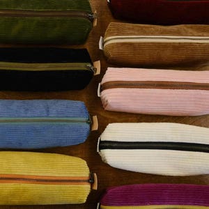 Può includere: Una collezione di astucci in velluto a coste in vari colori, tra cui verde, nero, blu, giallo, marrone, rosa, bianco, bordeaux e viola. Ogni astuccio presenta una chiusura con cerniera e una piccola linguetta in pelle. Disposti su una superficie di legno.