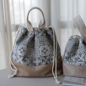 Peut inclure: Deux sacs &agrave; cordon dot&eacute;s d'un motif floral en dentelle gris fonc&eacute; et blanc sur la partie sup&eacute;rieure et d'une base en velours c&ocirc;tel&eacute; beige uni. Chaque sac poss&egrave;de une poign&eacute;e sup&eacute;rieure et des cordons blancs avec des n&oelig;uds.