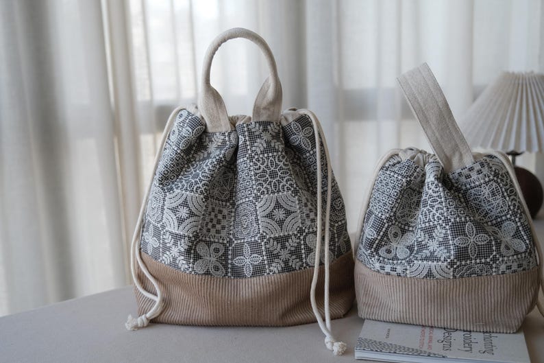 Peut inclure: Deux sacs &agrave; cordon dot&eacute;s d'un motif floral en dentelle gris fonc&eacute; et blanc sur la partie sup&eacute;rieure et d'une base en velours c&ocirc;tel&eacute; beige uni. Chaque sac poss&egrave;de une poign&eacute;e sup&eacute;rieure et des cordons blancs avec des n&oelig;uds.