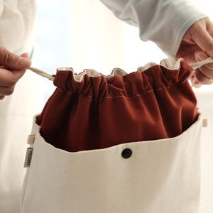 May include: A two-tone drawstring pouch featuring a cream-colored exterior with a single black snap button and a deep rust-colored fabric interior. The bag includes a cream-colored drawstring cord and a small rectangular tag on the side.