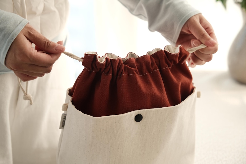 May include: A two-tone drawstring pouch featuring a cream-colored exterior with a single black snap button and a deep rust-colored fabric interior. The bag includes a cream-colored drawstring cord and a small rectangular tag on the side.