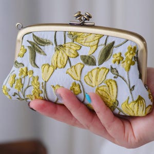 Puede incluir: Un pequeño bolso de mano azul claro con un marco de metal dorado. El bolso presenta un patrón floral bordado en amarillo y verde. El cierre es de diseño antiguo.