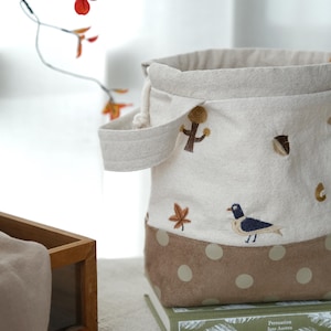 Op de afbeelding: Een cilindrische stoffen tas met trekkoord, voorzien van een geborduurde afbeelding van een vos, een vogel, een eikel en bladeren op het gebroken witte bovendeel. Het onderste deel is beige met lichtgekleurde stippen en heeft een zijhandvat.