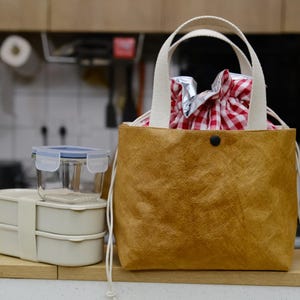 May include: A brown tote bag with white handles and a black button closure. Inside, a red and white checkered cloth holds a wrapped item. Beside the bag are two stacked beige lunch containers and a clear container with a blue lid.