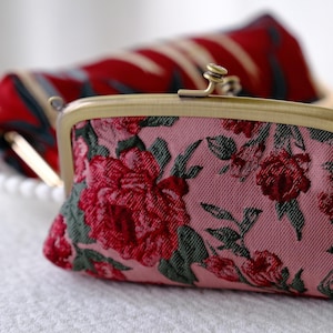 Puede incluir: Un monedero rosa floral con cierre de latón, con un estampado de rosas rojas y hojas verdes. Otro monedero con un estampado rojo y azul está ligeramente desenfocado en el fondo. Los monederos están sobre una superficie blanca.