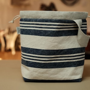 May include: A cylindrical storage bag with a drawstring closure. The bag features horizontal stripes in navy blue and off-white. The top and the small side loop are made of a solid off-white fabric. The bag is sitting on a light brown surface.