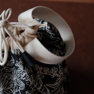 May include: Close-up of a drawstring bag with a black floral pattern on a dark background. The bag has a cream-coloured drawstring and a wide, off-white strap. The bag's interior is visible, showing a cream-coloured lining.