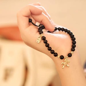 May include: Black beaded bracelet with gold-colored accents and a horse charm. The bracelet is made of round, dark beads and features a small tag and a horse pendant. The bracelet is held in a hand.