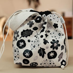 May include: A drawstring bag featuring a floral design in black and grey on a white backdrop. It has a light grey rim and white drawstring cords. The bag is made of fabric and is closed by pulling the cords.