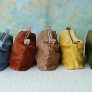 Puede incluir: Cinco estuches rectangulares con cremallera hechos de tela de pana en azul, óxido, bronceado, amarillo y verde oliva. Cada estuche cuenta con un cierre de cremallera superior con una pequeña lengüeta de tela estampada en el tirador, con textura acanalada.