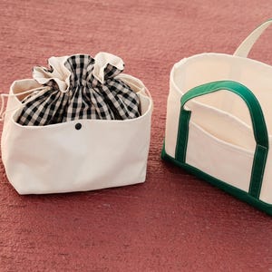 May include: Two canvas tote bags. One is a white drawstring bag with a black and white checkered interior. The other is a white tote bag with green handles and trim. Both bags are on a red surface.